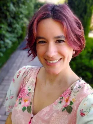 lined with green hedges. She has hair cut into a wavy, jaw-length bob and dyed pink. Her top is a floral patterned pink V-neck with white, puffy capped sleeves. She is smiling directly at the camera.